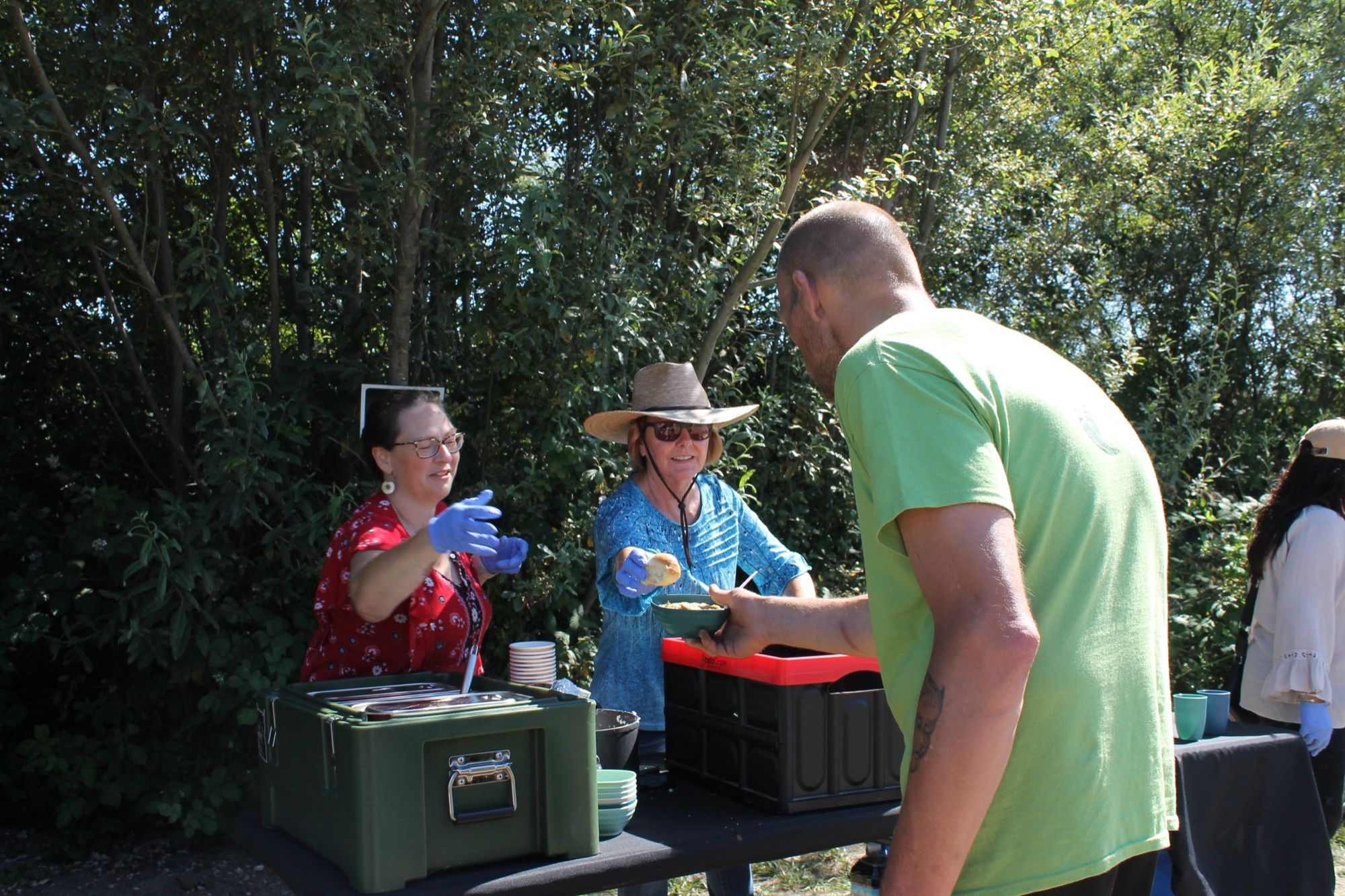 A volunteer in a cowboy hat hands a bowl of soup across the table to a man in a green shirt