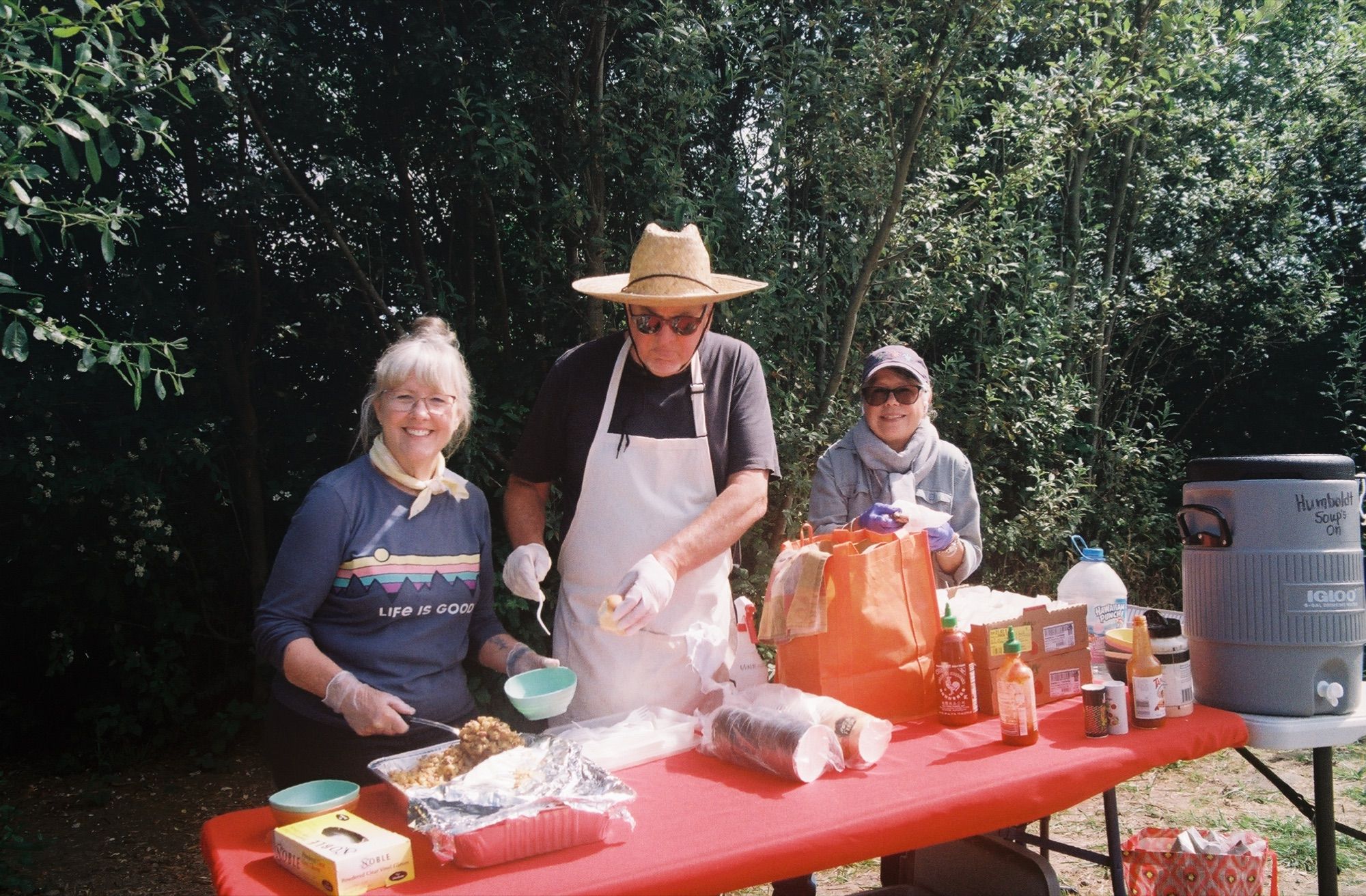 Three volunteers in aprons and gloves behind a red table of food, in the early days of serving on-site