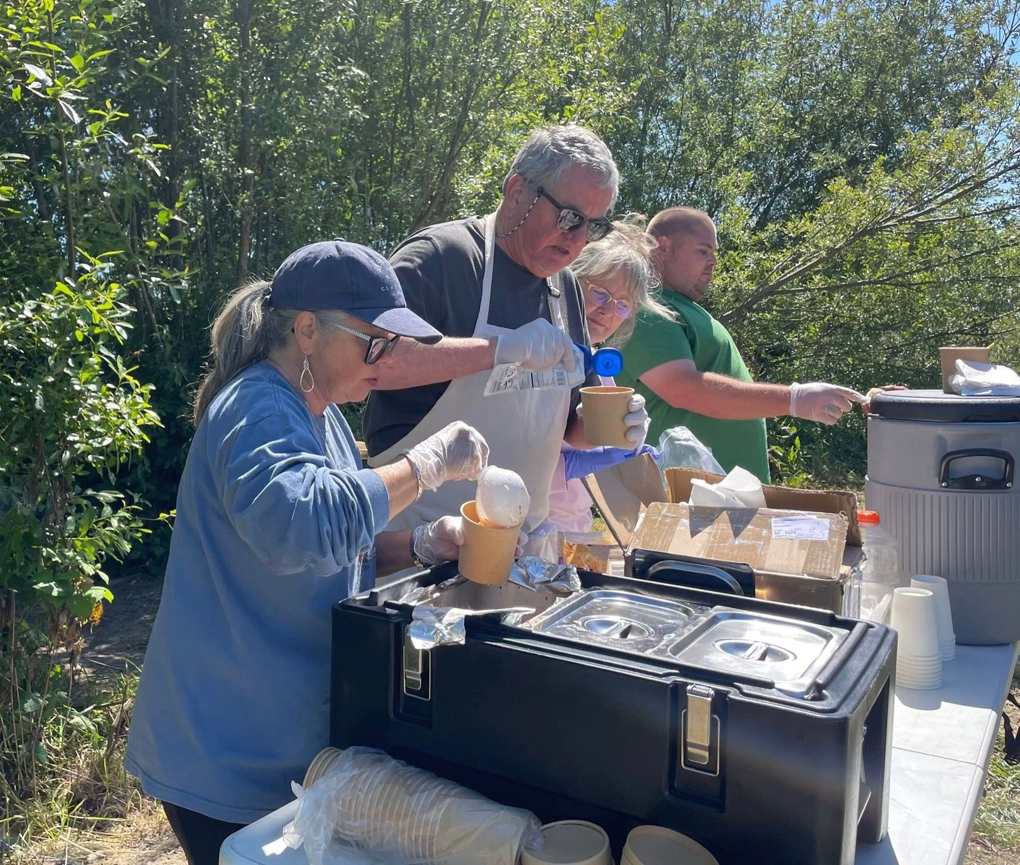 Volunteers in aprons ladling soup into paper cups at an outdoor table